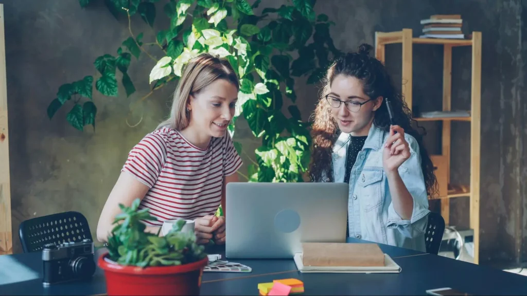 Two women collaborate at a table with a laptop in a plant-filled workspace, discussing something.